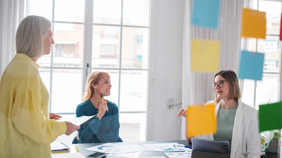 Three professionals in a bright office engaged in a discussion around a table with documents and charts, with colorful sticky notes on glass in the foreground.