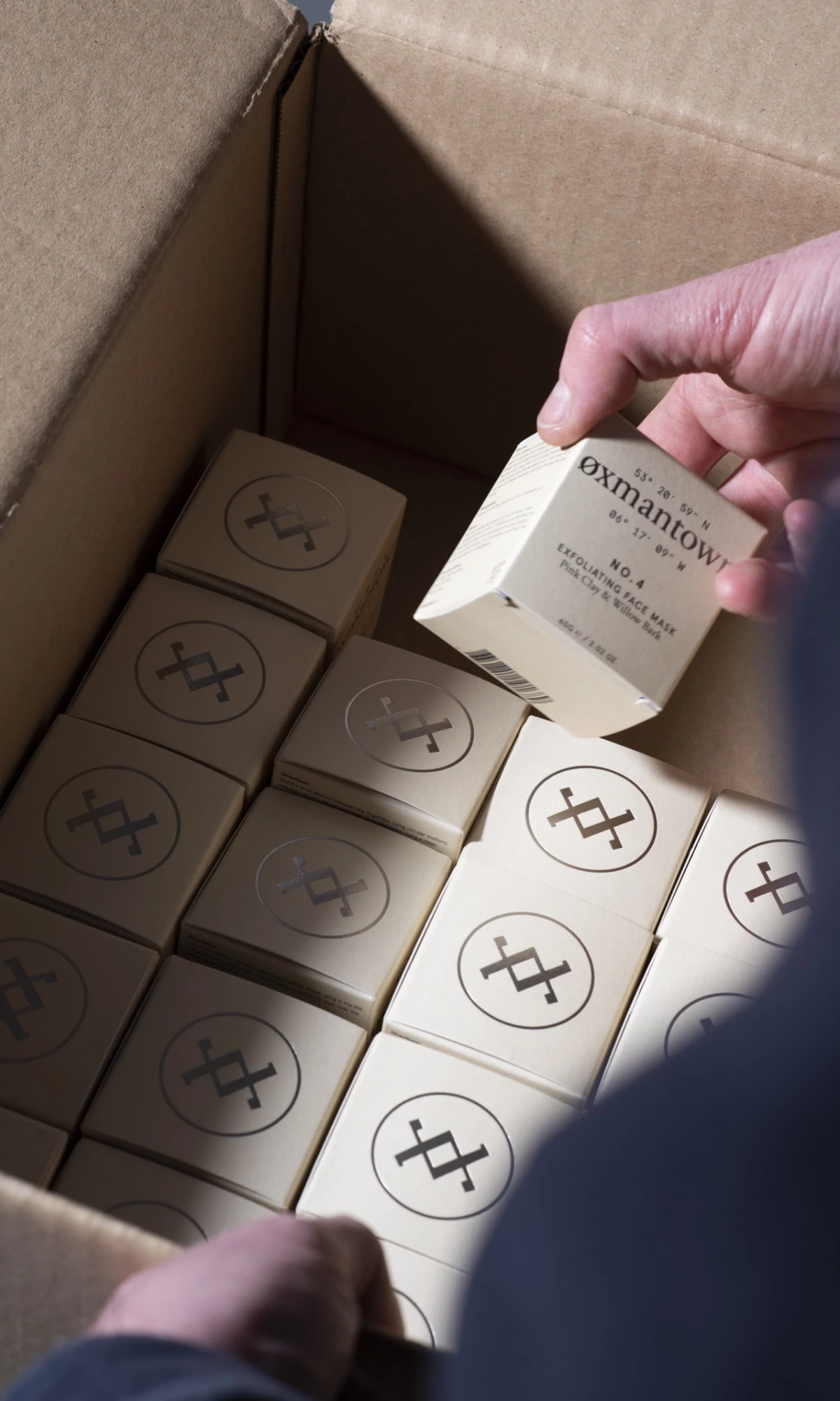 Hand placing boxes of cosmetics into a large cardboard box featuring oxmantown skincare branding