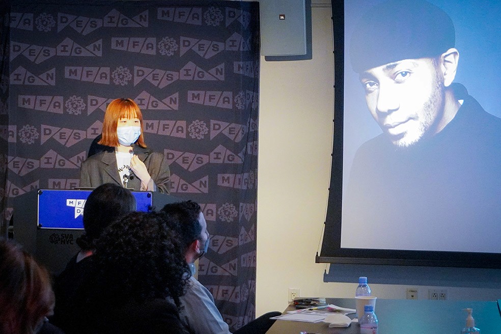 A woman is giving a speech to the public, while on the right side of the wall is a projection of a black and white portrait of a man with a hat