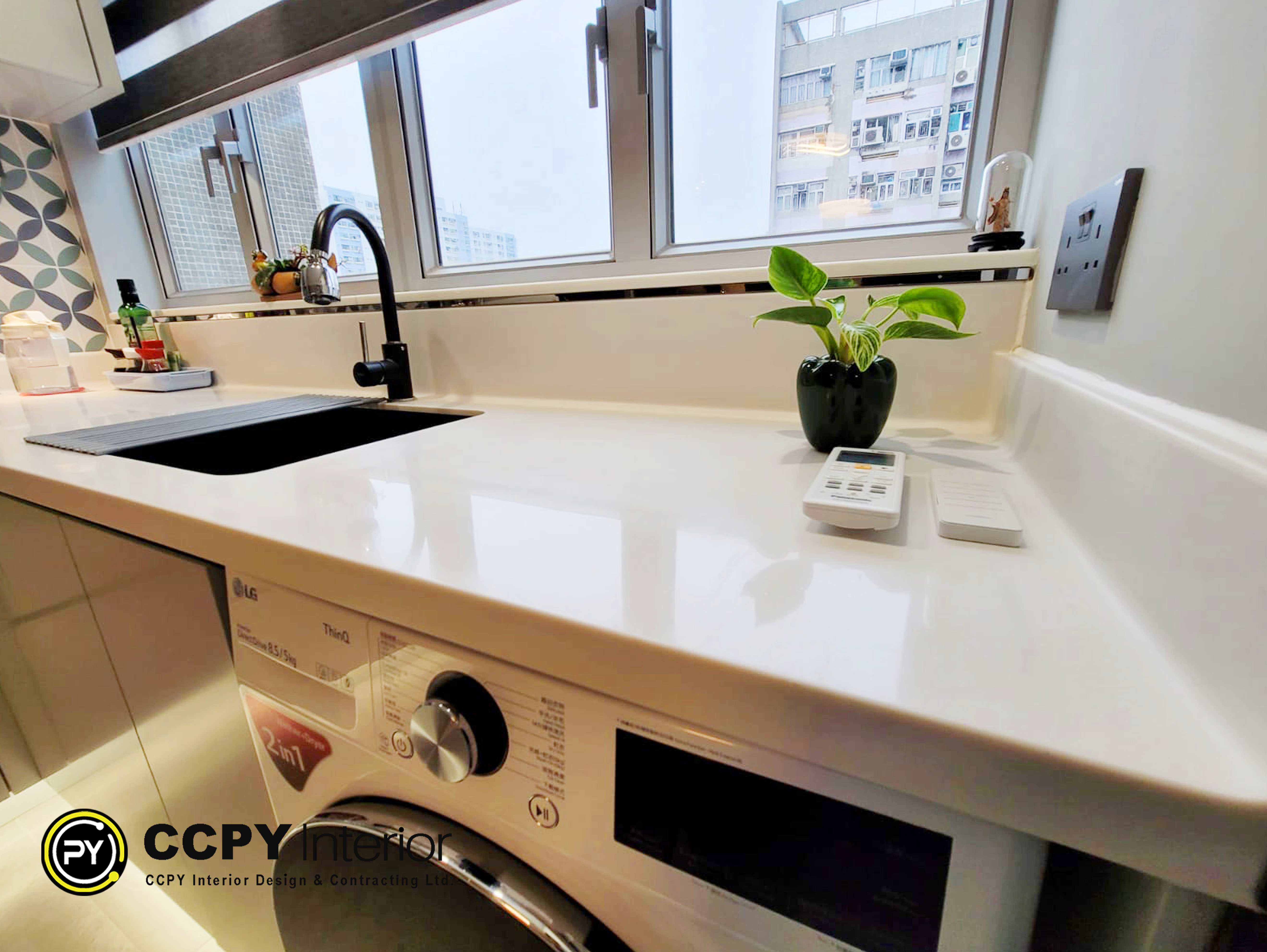 The selected region shows a bright, modern open-plan kitchen counter that doubles as a utility zone featuring a built-in washing machine, a black single-bowl sink, a matte black faucet, and various decorative and functional countertop items.Design and Material DetailsBased on the design notes provided in the document, here are the explicit details regarding this specific area:Kitchen Layout: Designed as an open-plan kitchen to maximize spatial harmony and day-to-day functionality.Cabinetry: Gray hand-painted lacquer solid timber plywood cabinets that provide a clean, modern aesthetic.Backsplash Tiles: British-style backsplash tiles arranged in a distinct geometric pattern on the left wall.Appliances: A front-loading washing machine integrated directly underneath the solid white countertop.