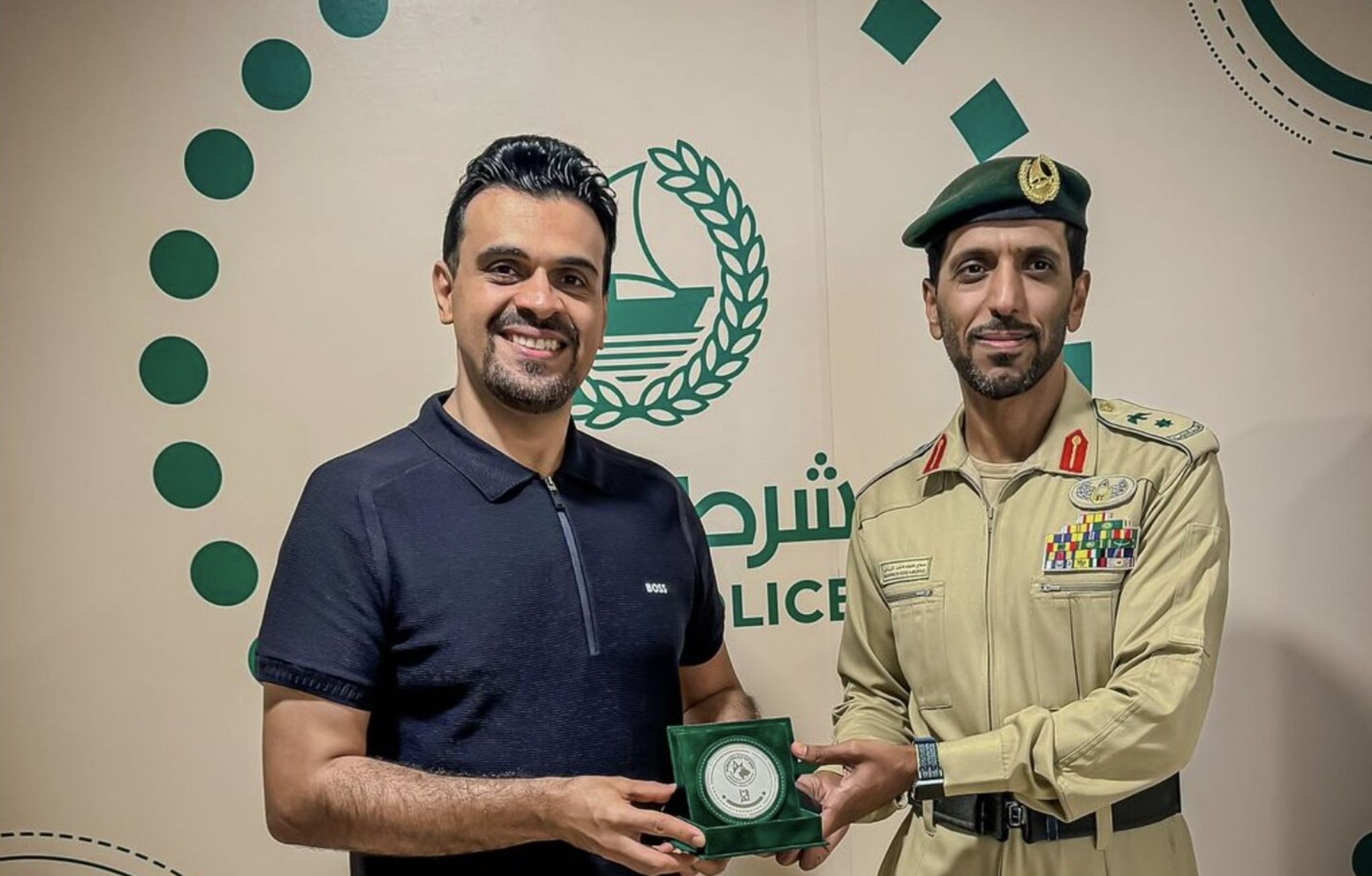 Dr. Soheyl Simaei (left), wearing a dark polo shirt, smiles while receiving a commemorative plaque from Lt. Colonel Salah Khalifa Al Mazrooei (right), dressed in a decorated beige police uniform with a green beret. They are standing in front of a wall displaying the Dubai Police emblem.