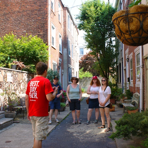 A tour guide talks to four people in an alley lined with brick buildings and greenery.