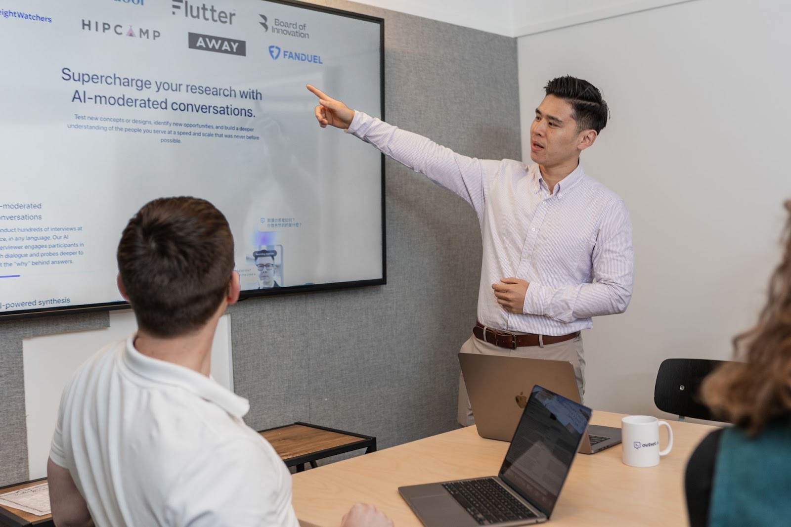 Person standing and pointing to a large presentation screen while a small group watches; laptops and mugs are on the table