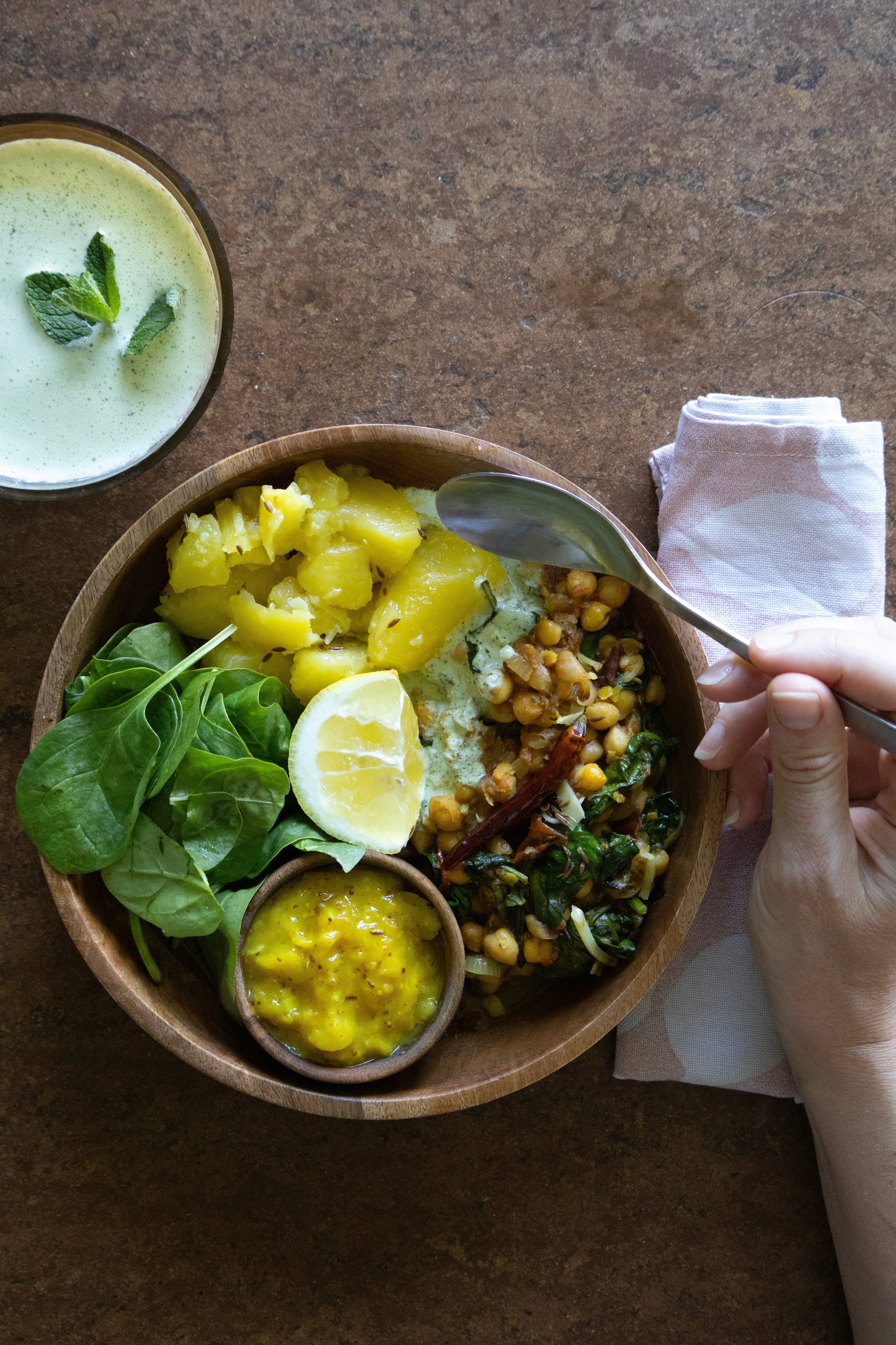 Eine Hand hält einen Löffel über einer mit Schüssel mit Kichererbsen-Kokos-Curry, Kartoffeln, Mango-Dip