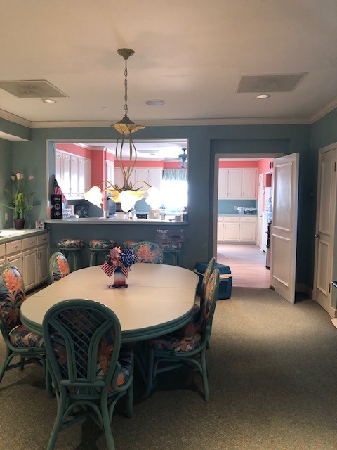 A coastal-themed dining room features a large, white oval table with teal, wicker chairs, adorned with floral cushions, set under a unique, leaf-shaped chandelier, with a view into an adjacent kitchen with light cabinets and a coral accent wall in the background.