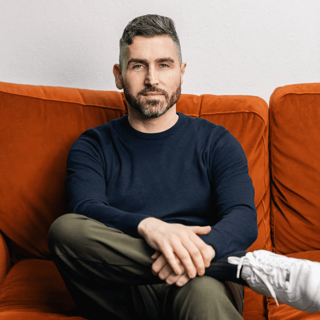 Man in dark sweater on orange couch, looking at camera.