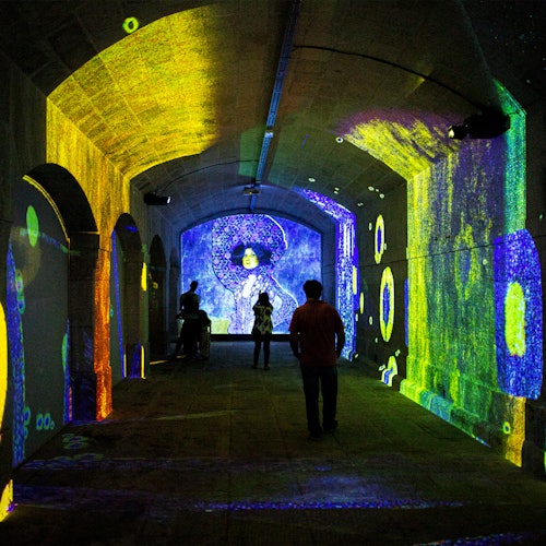 As pessoas caminham por um túnel iluminado com projeções coloridas e abstratas nas paredes e no teto.