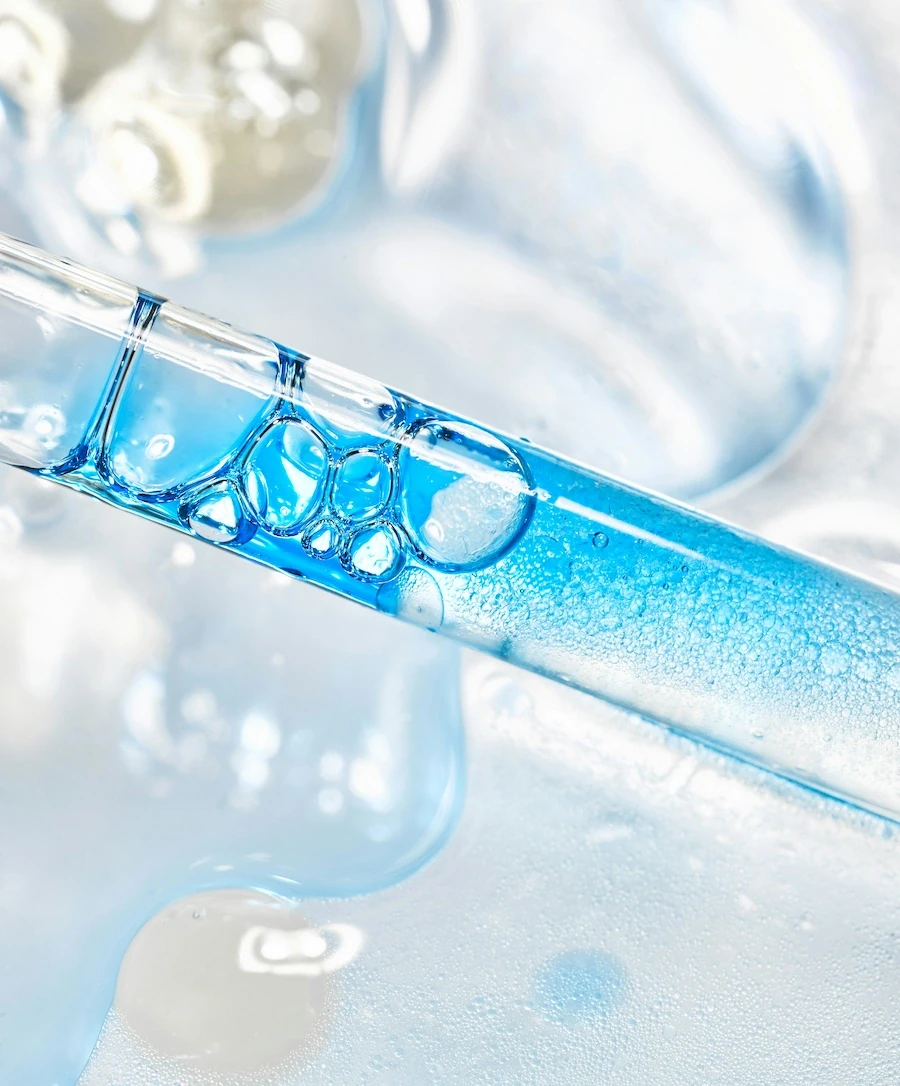 Macro photograph of a glass test tube containing vivid blue liquid with bubbles, resting on a reflective surface surrounded by laboratory glassware; representing in-vitro research and purity analysis.