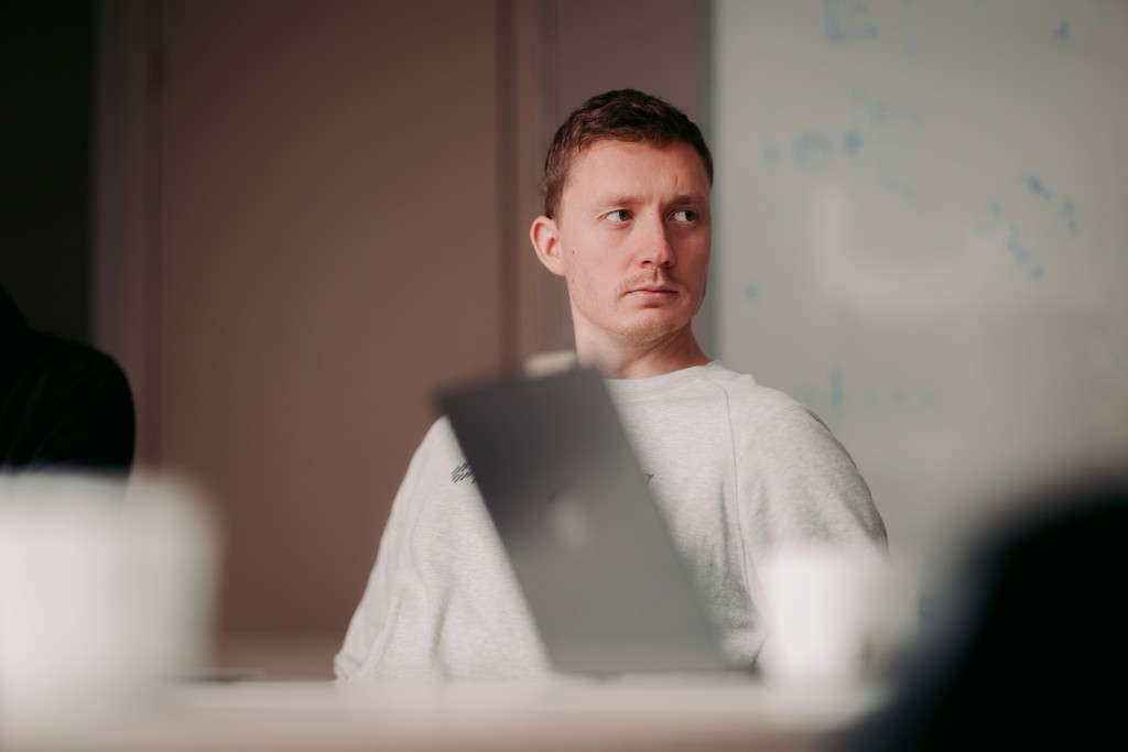 A person in a light sweater is sitting at a conference table with a laptop in front, appearing thoughtful and looking to the side, in a dimly lit office setting.