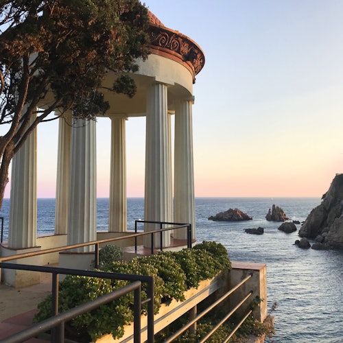 一个面向海洋的柱式亭子在夕阳下，前景有岩石露头和郁郁葱葱的绿色植物。