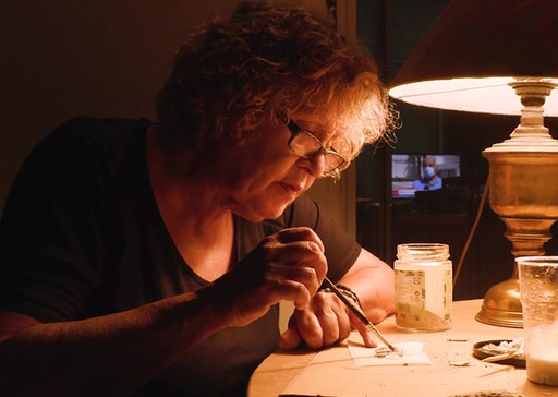 Η Ρένα Παπασπύρου εργάζεται σε τραπέζι με λάμπα, κρατώντας πινέλο.