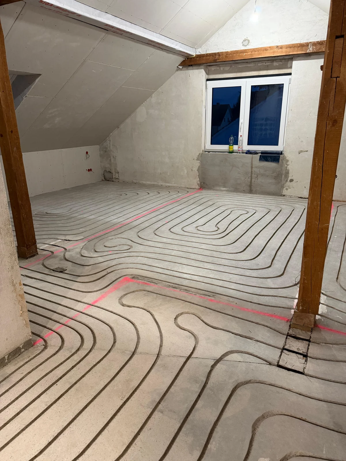 Gefräste Heizkanäle im Estrich eines Dachraums mit sichtbaren Holzbalken und Fenster.