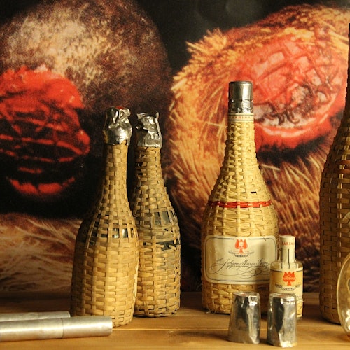 Empat botol yang dibungkus anyaman, termasuk satu botol lebih besar dengan label, dan satu botol miniatur di permukaan kayu dengan latar belakang bertekstur.