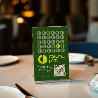 Close-up of a "Casual Dot" table tent sign with a QR code, on a white table. The sign has a green background.