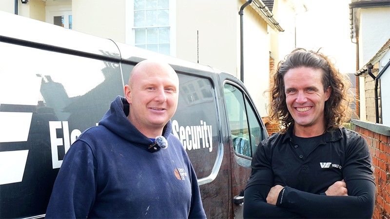 Two men stand smiling in front of a van, one in a hoodie and the other in a black shirt, against a suburban backdrop.