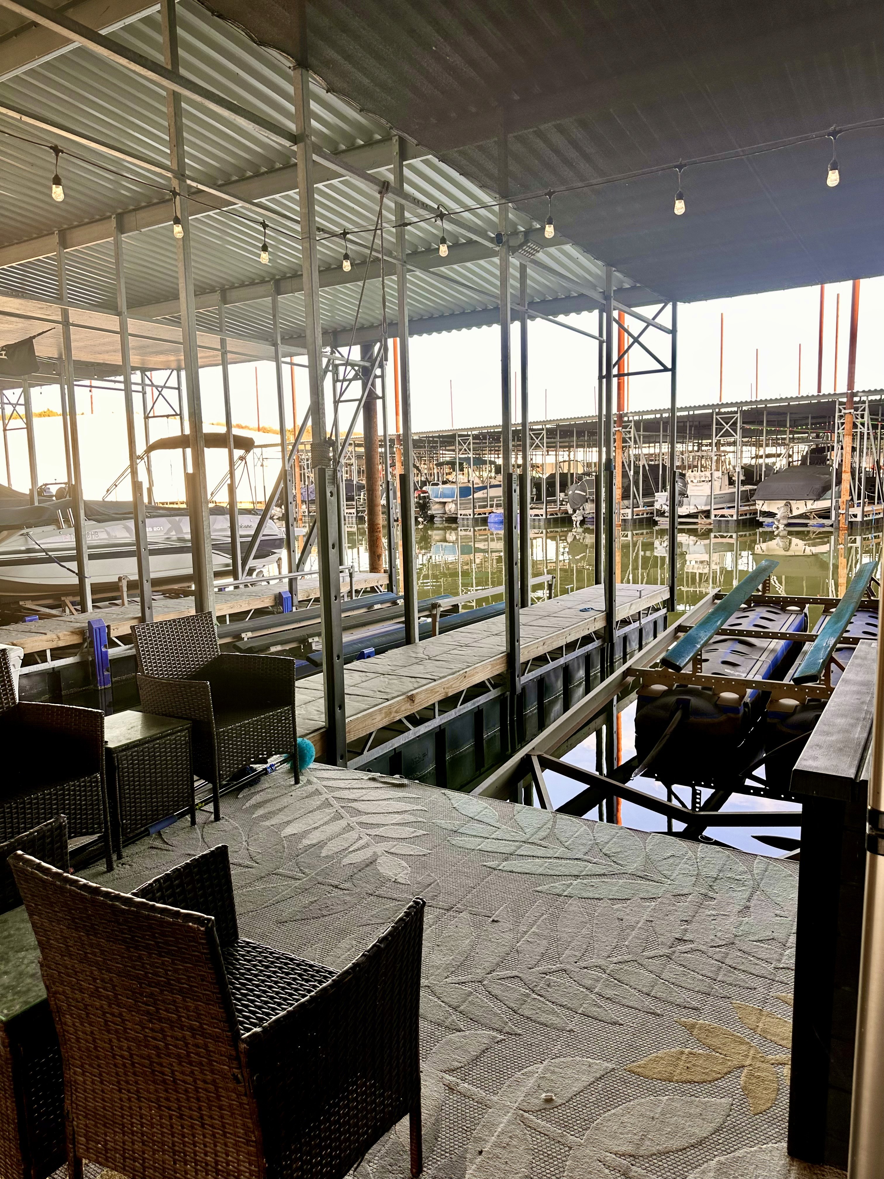 A modern marina features neatly arranged slips and a spacious deck with wicker chairs, providing a relaxing view of various docked boats and the calm waters under a corrugated metal roof.