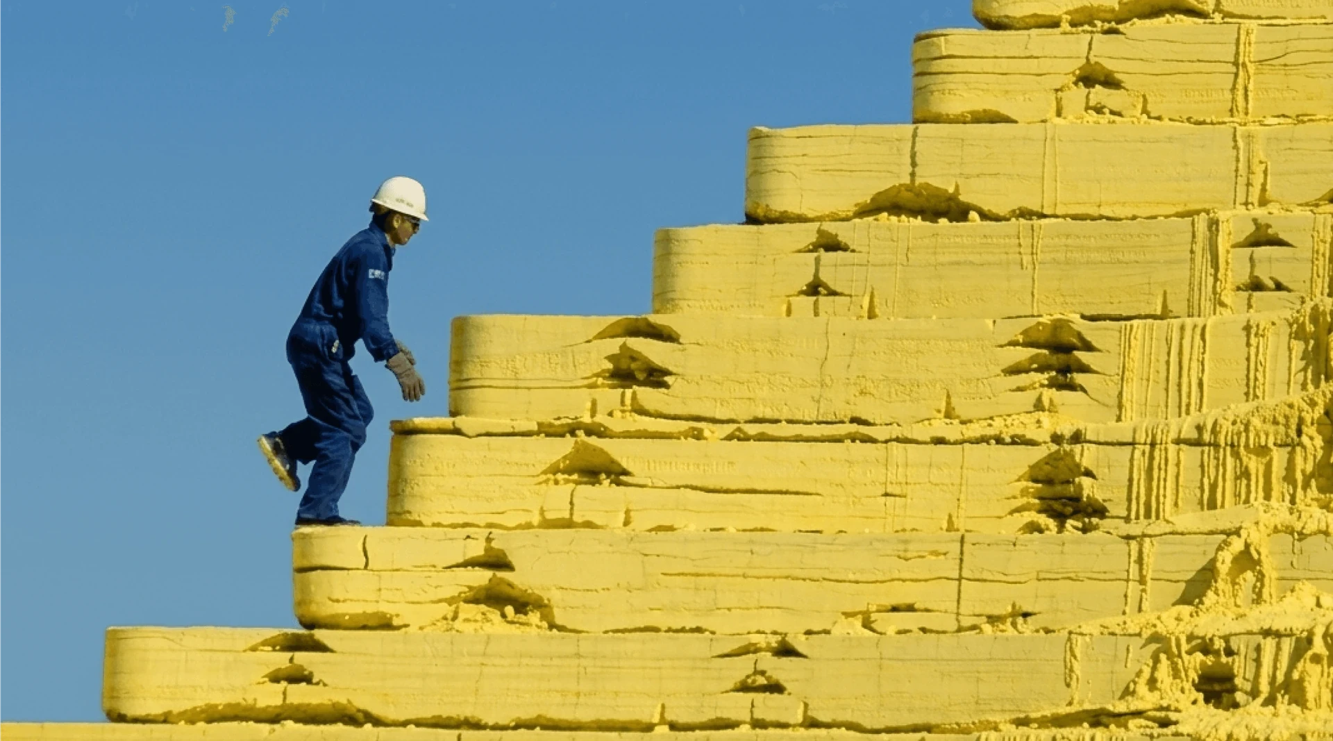 A worker climbing a large mound of yellow sulfur under a blue sky