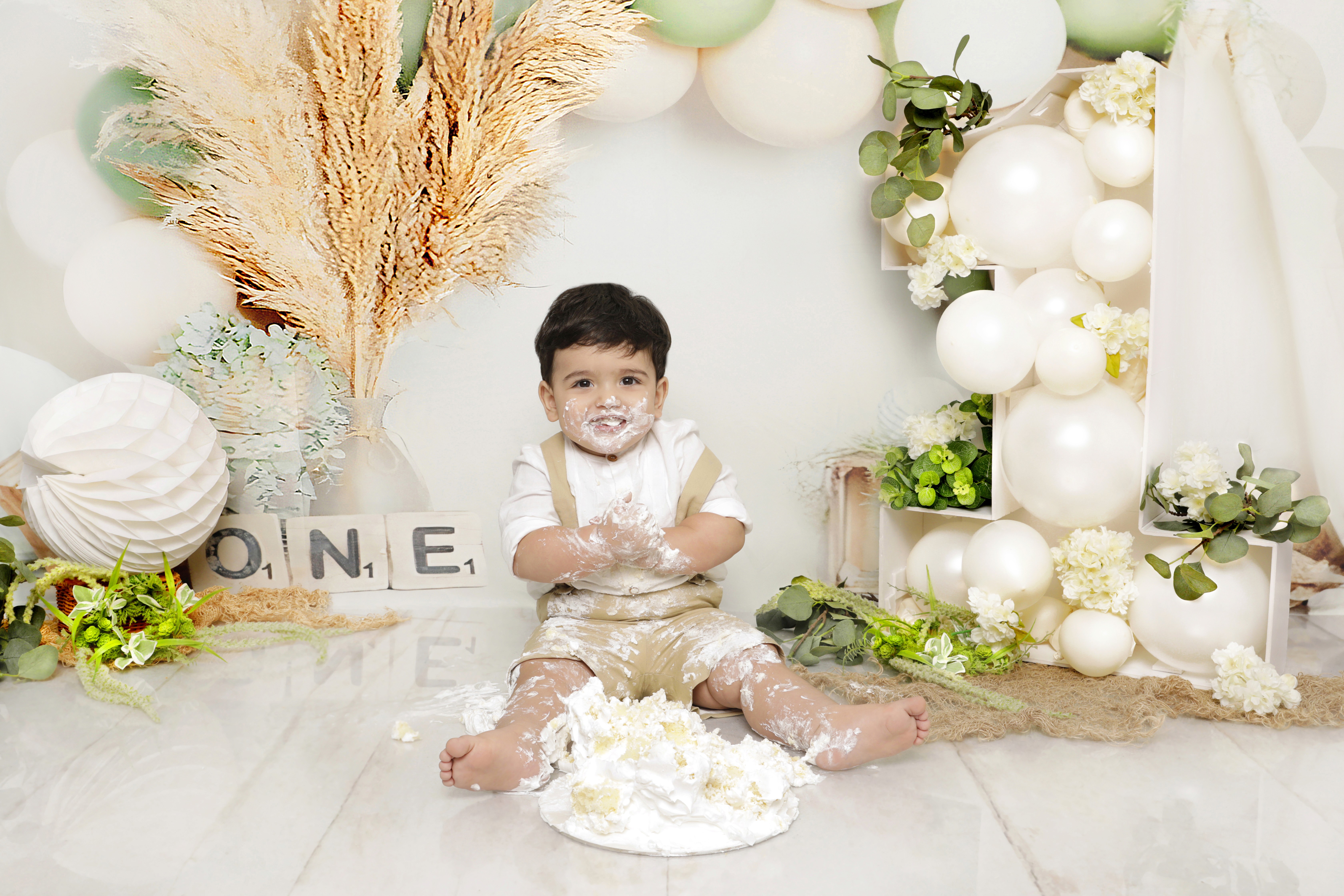 A baby is sitting in a white cake smash setup with balloons, greenery, and “ONE” blocks for a monthly baby photo. He is covered in white frosting. 