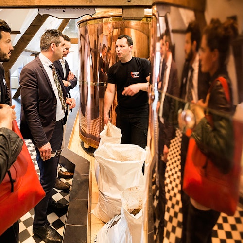 En gruppe mennesker i forretningsantrekk lytter til en mann som forklarer i et rom med industrielle kar og sekker på et rutete gulv.