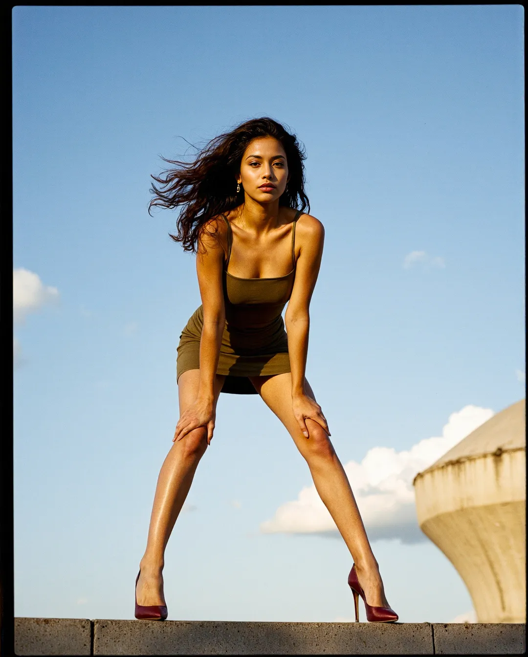 Fashion editorial photograph featuring a model in olive dress and burgundy heels posing confidently against bright blue sky