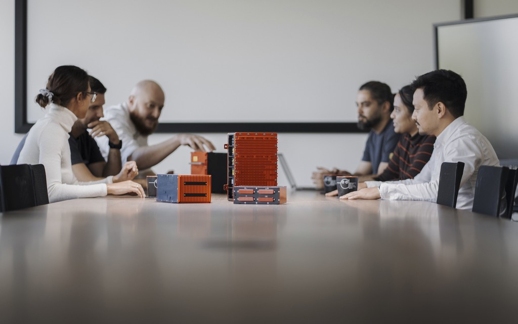 A group of six people engaged in a discussion around a conference table in a bright meeting room. On the table are products of AEE