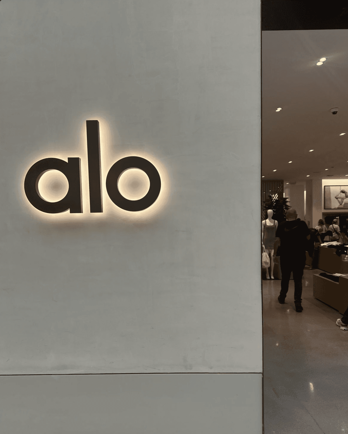 Illuminated “alo” storefront sign mounted on a white wall at the entrance of an Alo Yoga retail store.