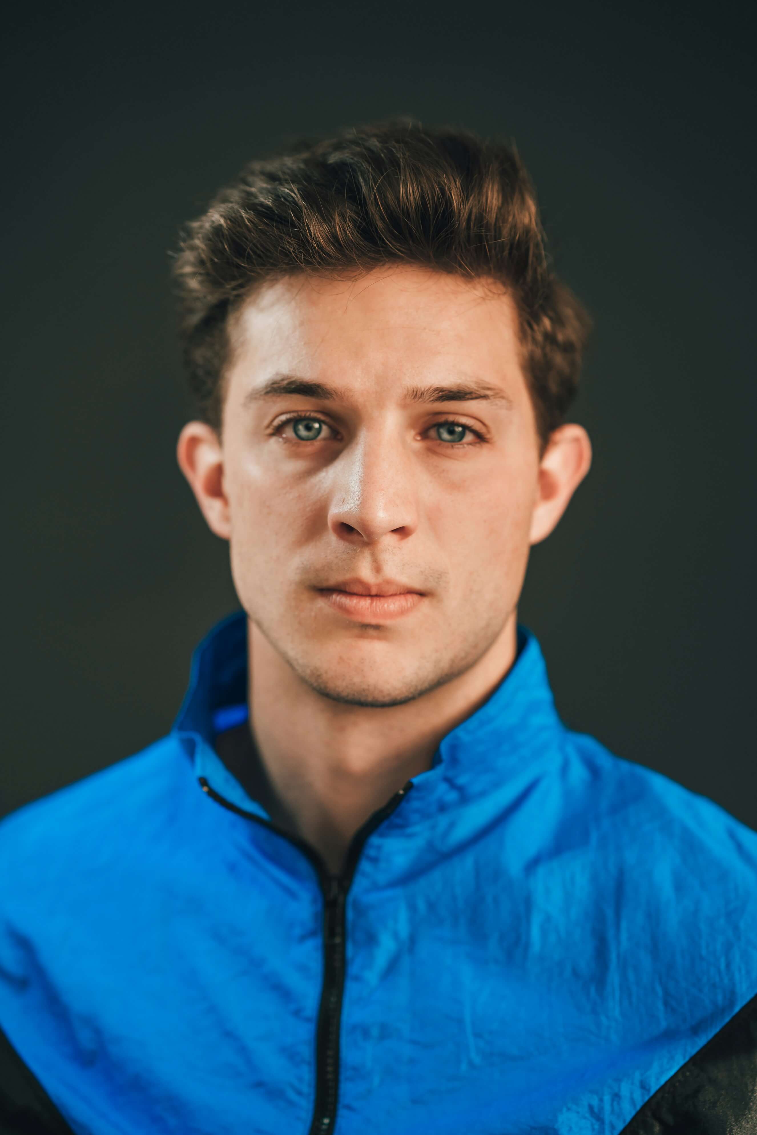 Portrait of a young man with brown hair and blue eyes, wearing a blue jacket, looking directly at the camera.