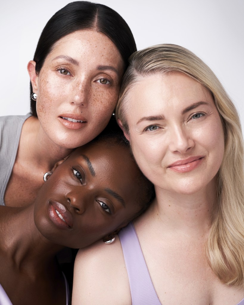 Three women of diverse backgrounds pose closely together, smiling and showcasing their natural beauty. The woman on the left has long, dark hair with freckles, the woman in the middle has a dark complexion, and the woman on the right has blonde hair. They are wearing soft-toned clothing against a neutral background.
