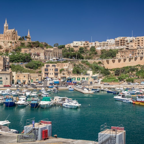 A harbor with various boats docked, surrounded by buildings and a hilltop church under a clear blue sky.