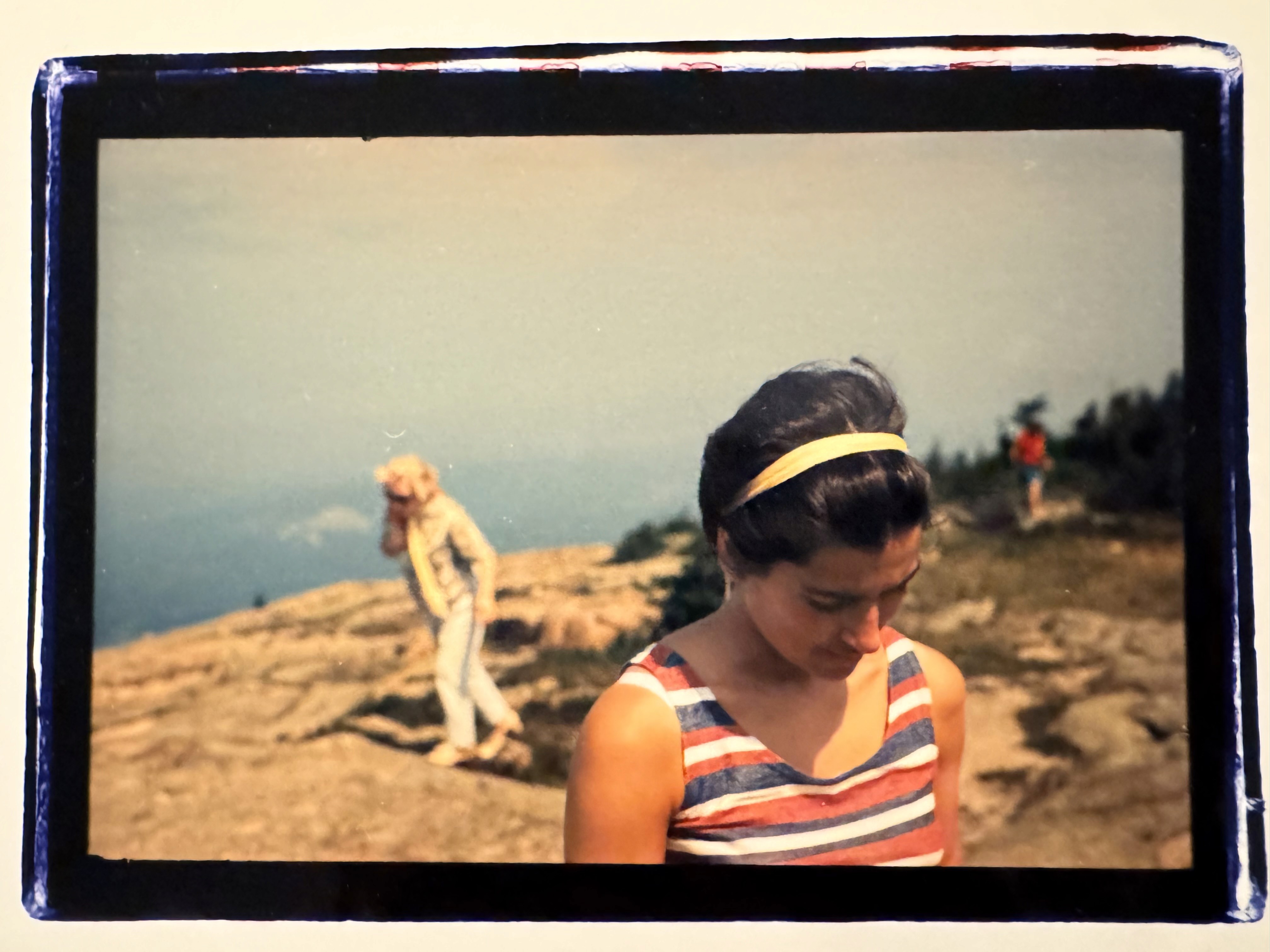 A young woman wearing a yellow headband in her short dark brown hair and a red white and blue striped tank top, looks down out of frame, a landscape behind her and an older woman walking at a distance with a similar posture.