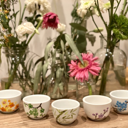 五个装饰性的陶瓷碗,上面有花卉图案,陈列在木制表面,前面摆放着装在玻璃罐中的各种花朵。