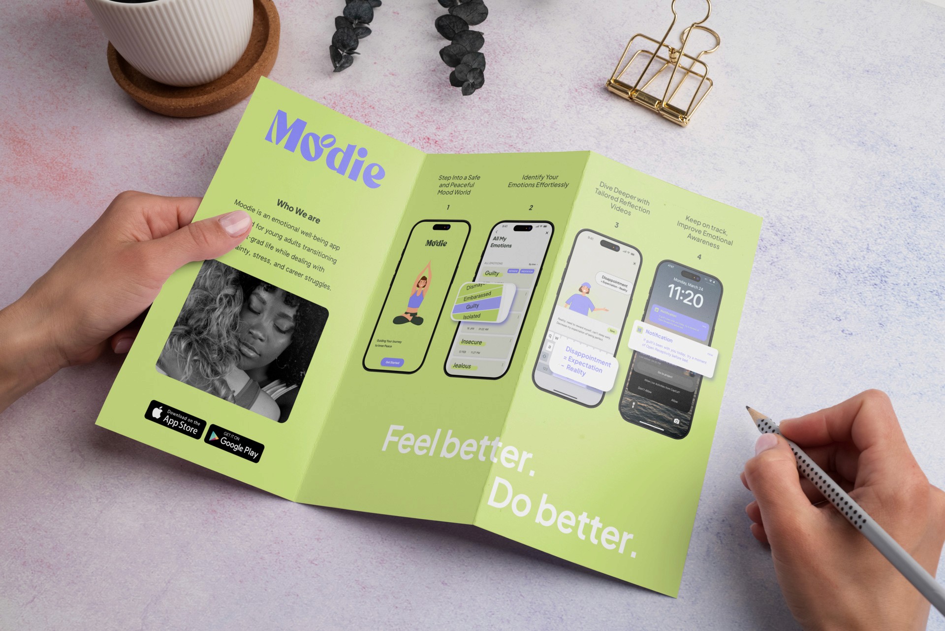 Two hands hold open a brochure titled “Moodie,” displaying four smartphone screens showcasing different apps and features. The brochure’s text reads “Feel better. Do better.” and includes descriptions like “Who we are,” “What we do,” and “How we do it.” A coffee cup, coaster, wire organizer, and dried flower petals are visible in the background.