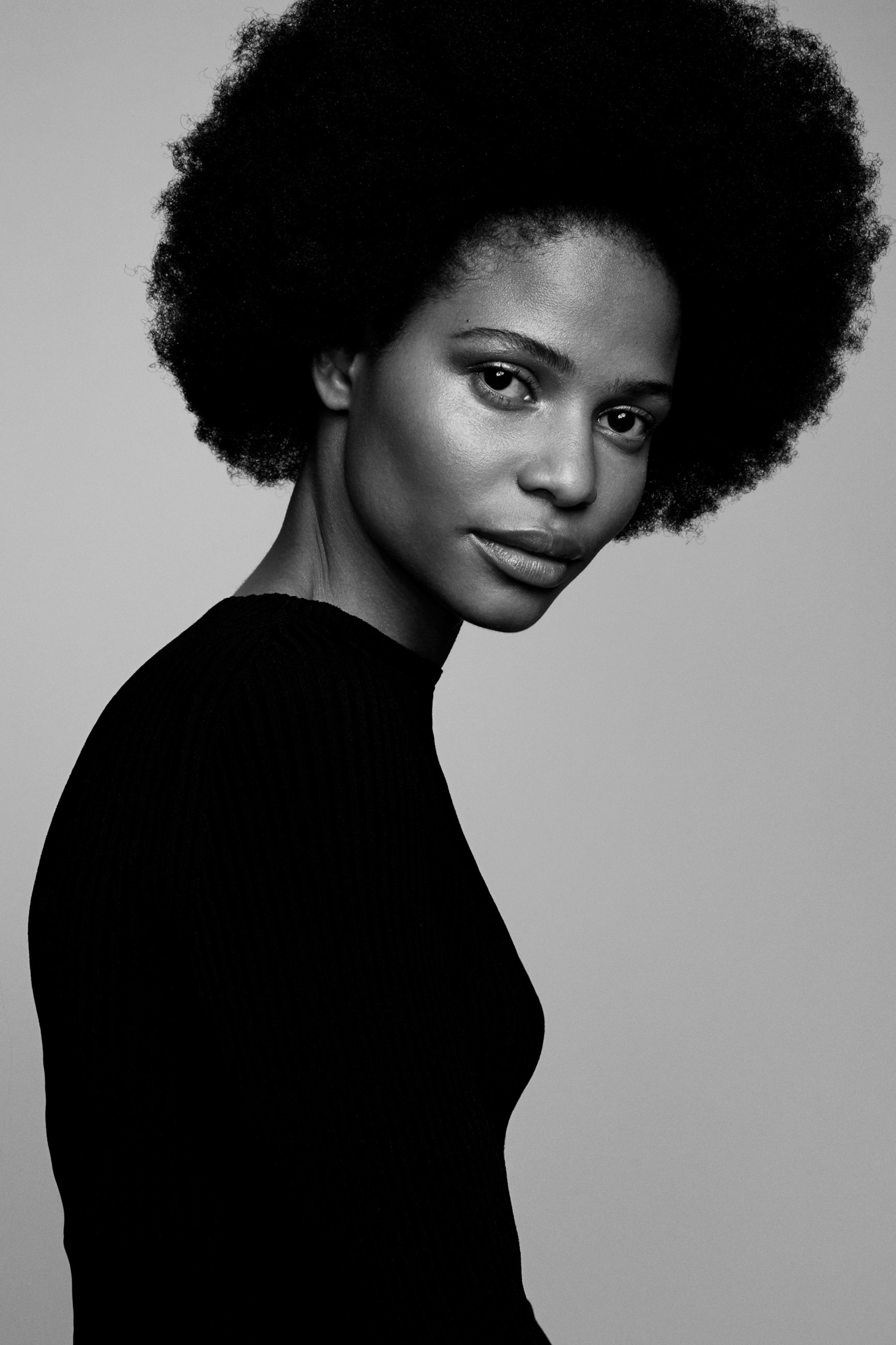 Classic black and white fashion portrait of a black model with a natural afro hairstyle wearing a tight black knitted turtleneck top so her body is silhouetted against a minimal light grey background.