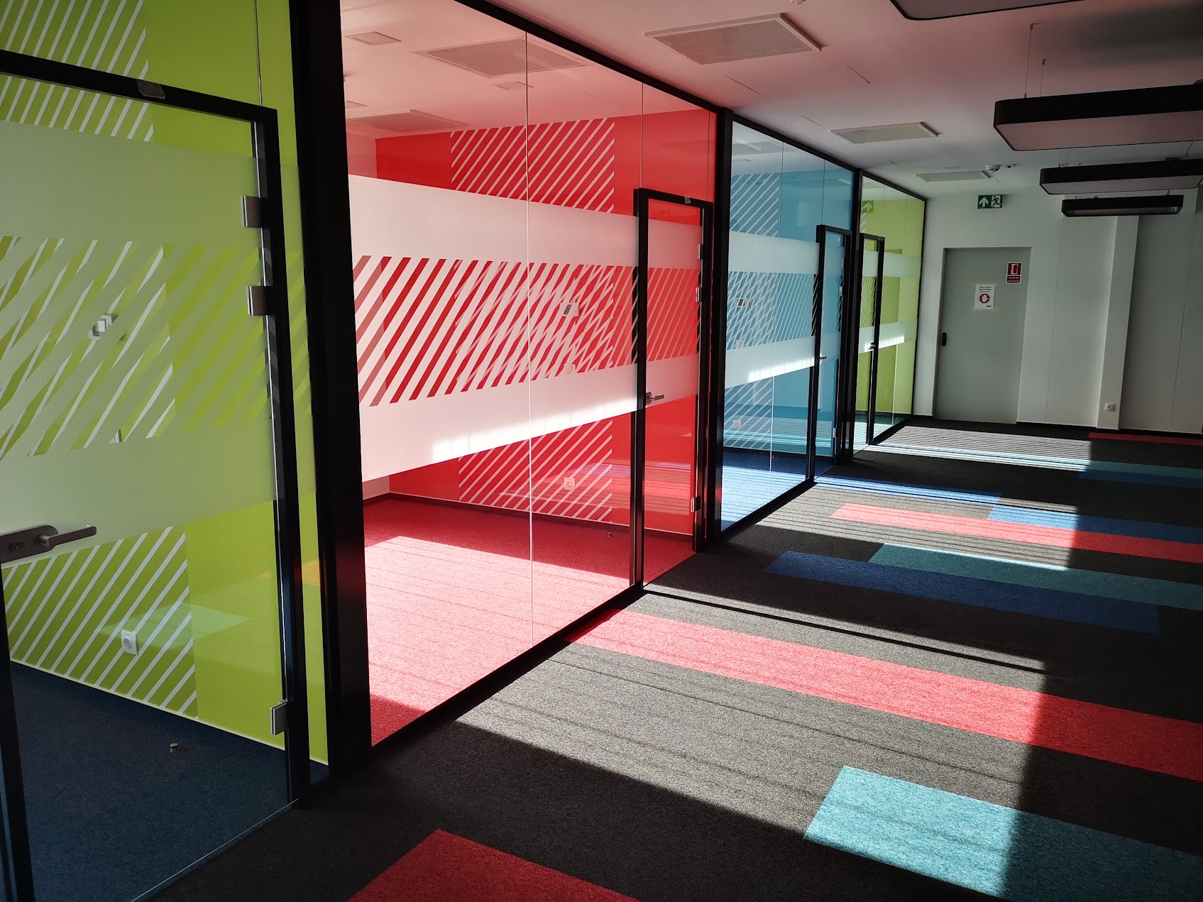 Modern irodaépület folyosója élénk zöld, piros és kék színű falakkal, teliüveges irodákkal amelyeken átlósan csíkozott tejüveg fólia biztosítja a szekciók belátásvédelmét. Prémium irodai dekorfóliázás és térelválasztás.