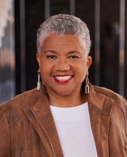 Smiling person with short gray hair, wearing a brown leather jacket over a white shirt, in an outdoor setting.