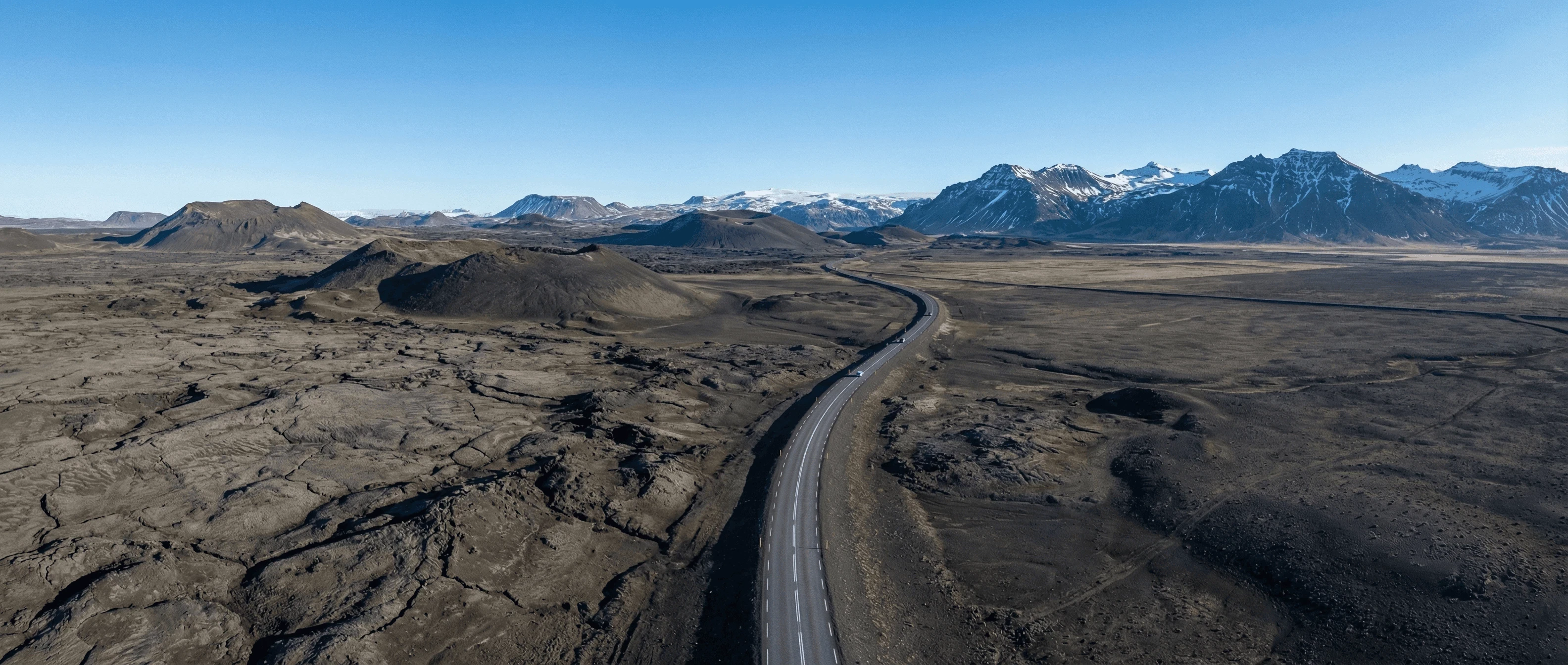 A paved road winding through a vast, barren black lava landscape towards distant mountains under a blue sky.