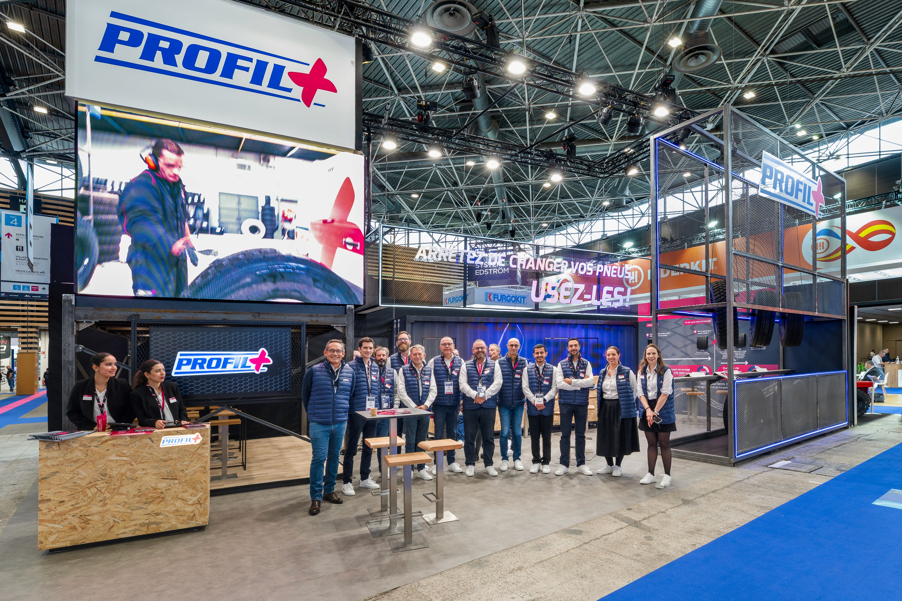 Stand Profil+ Solutrans Lyon 2026 conçu par Expace – vue frontale animée du stand avec visiteurs professionnels, écrans bleu électrique et rose et structure conteneur industrielle