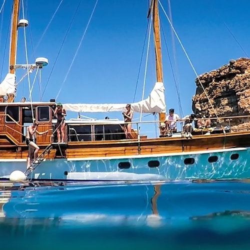 People aboard a wooden sailboat, anchored near a rocky coastline under a clear blue sky, some preparing to dive into the water.