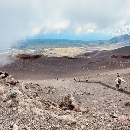 People hiking down a rocky, volcanic slope with a distant view of plains and mountains under a cloudy sky.
