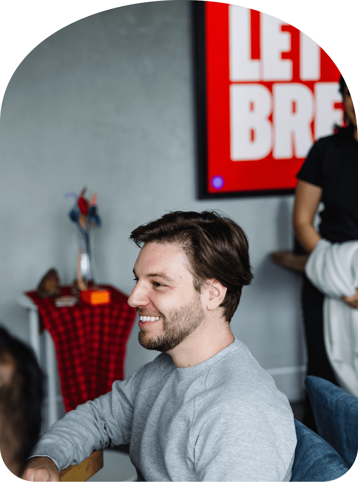 Man smiling while seated at a table in an office setting, with a bold red sign partially visible in the background.