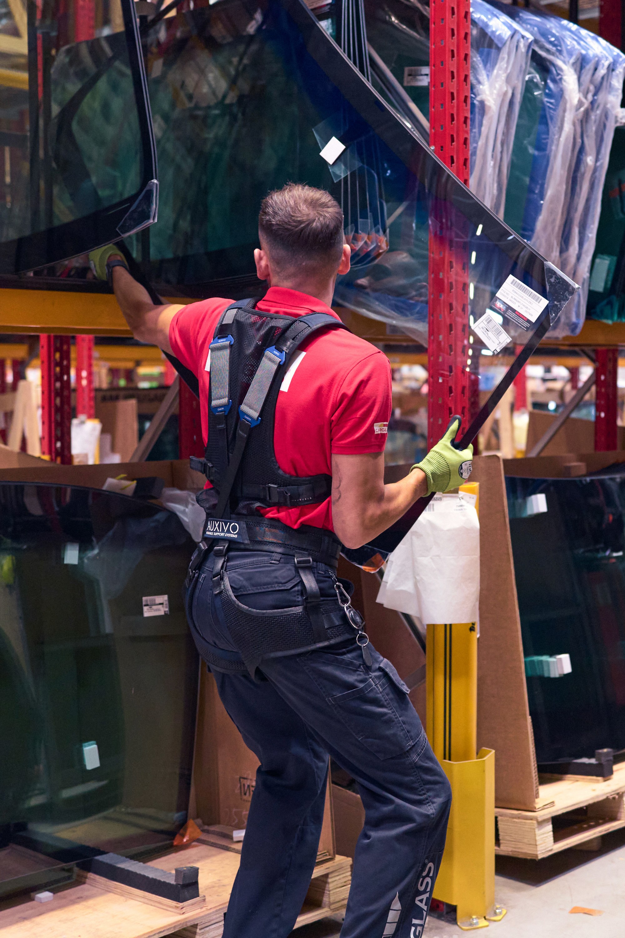 Travailleur en entrepôt portant l'exosquelette LiftSuit Auxivo noir, manipulant de grandes vitres dans un entrepôt de distribution, rayonnages industriels rouges en arrière-plan.