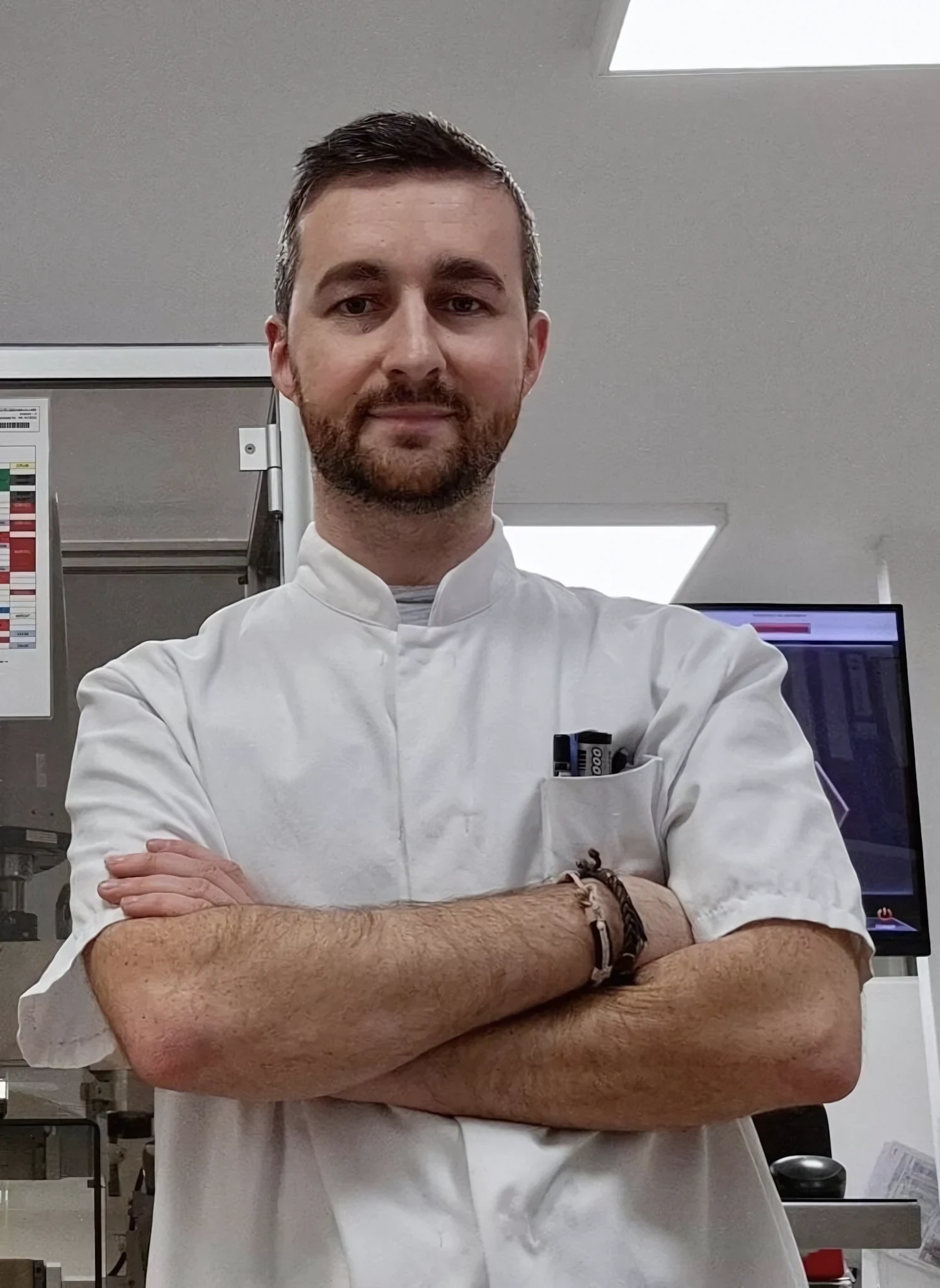 A smiling healthcare professional with short hair is wearing a white lab coat and a stethoscope around their neck, standing in a well-lit room with a green plant in the background.