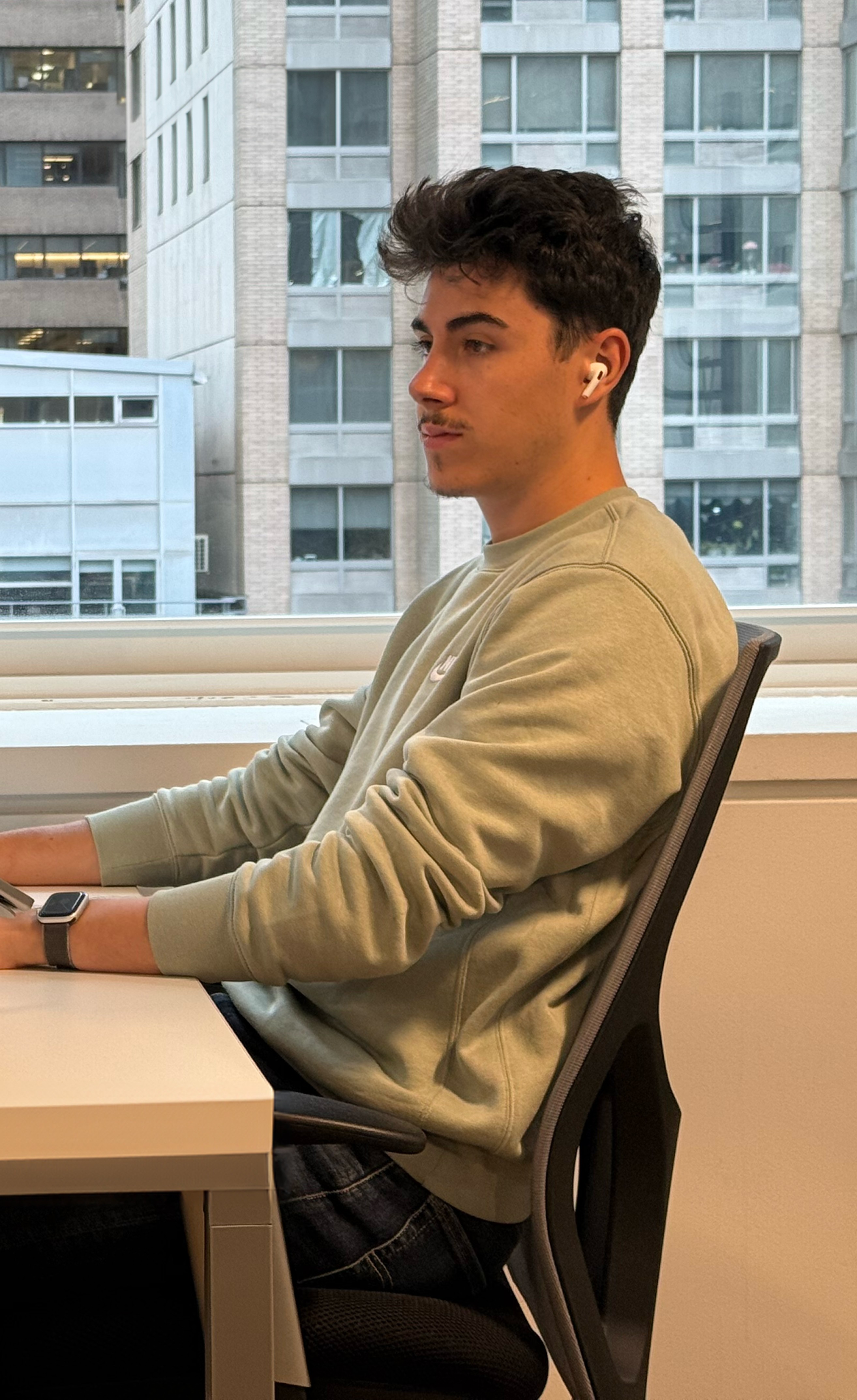 Freelance assis à son bureau dans un open space moderne, travaillant sur un MacBook sur support ergonomique, avec vue sur des immeubles urbains.