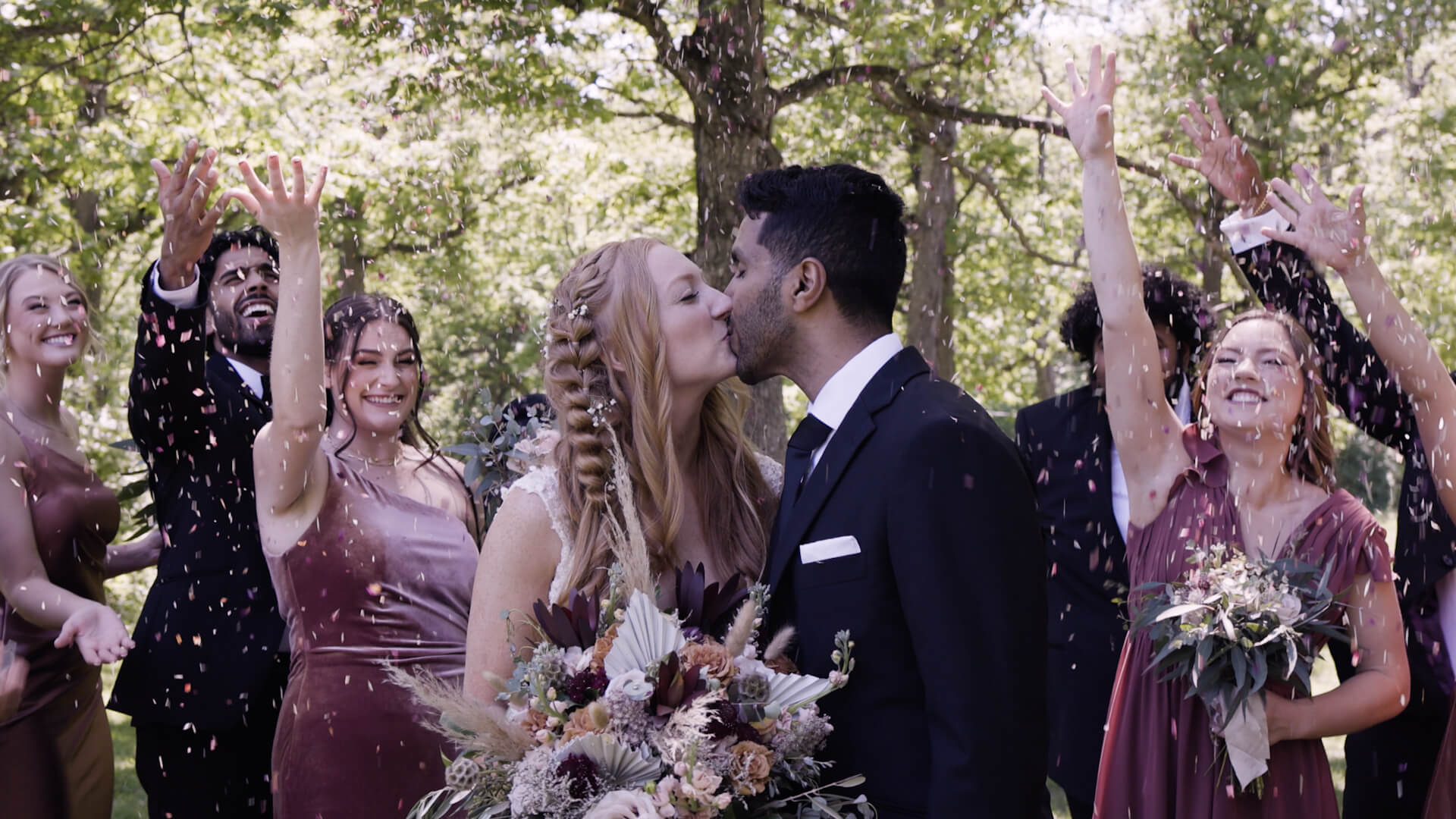 A bride and groom kiss while bridesmaids and groomsmen shower them with confetti