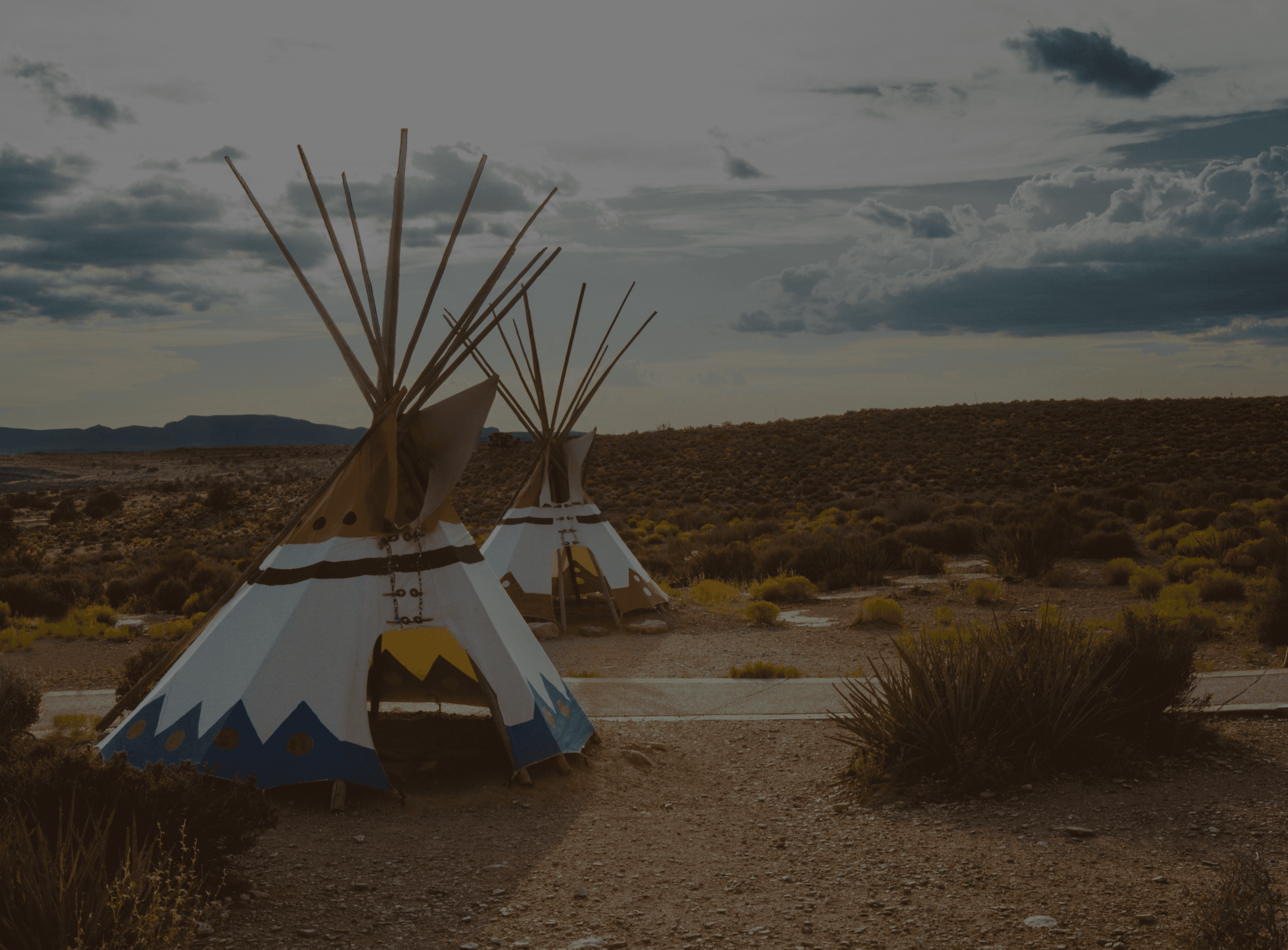 Two tipis stand in a desert landscape under a cloudy sky, surrounded by sparse vegetation.