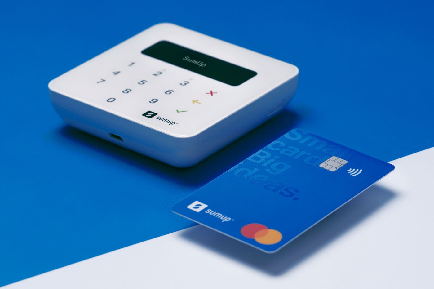 A white, square-shaped credit card reader with a digital display and numeric keypad is placed next to a blue credit card featuring contactless payment symbols, on a vibrant blue surface.