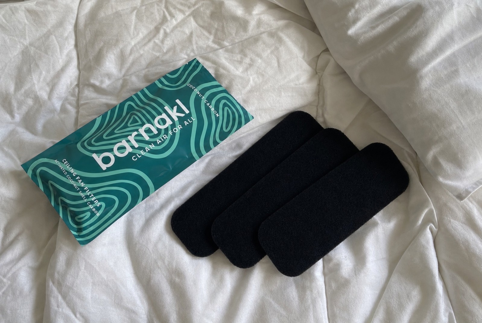 The Barnakl pouch and three matte black filter pads on white bed linens in morning light.