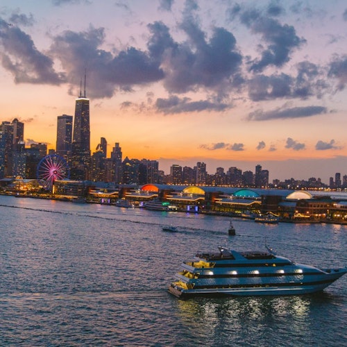 Isang bangka ang nagkrus sa ilog sa dapit-hapon na may tanawin ng lungsod sa likuran, tampok ang isang Ferris wheel at nagniningning na mga gusali.