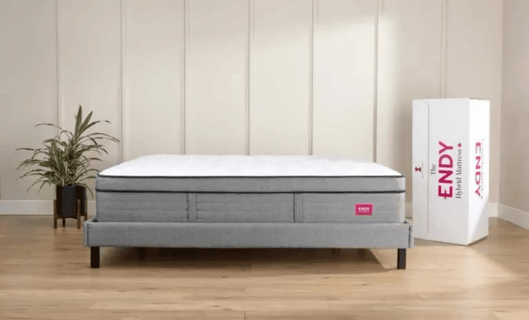 An Endy Hybrid Mattress shown on a grey upholstered bed frame against a light paneled wall. A standing white Endy box is next to the bed, used for comparison in the newcomer mattress-in-a-box guide.