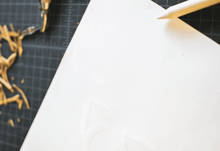 A white sheet of paper lies on a dark gridded surface. A pencil rests on the upper right corner of the paper. Near the left edge, a pile of thin, light-colored strips is scattered across the surface.